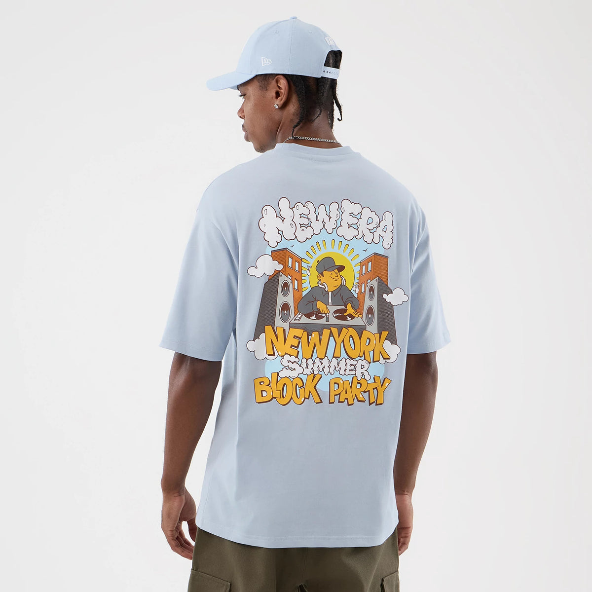 New Era Fun Block Party Graphic Pastel Blue Oversized T-Shirt, back view on a model styled with a matching cap, featuring a cartoon DJ scene with New Era and New York Summer Block Party text in lettering across the back.