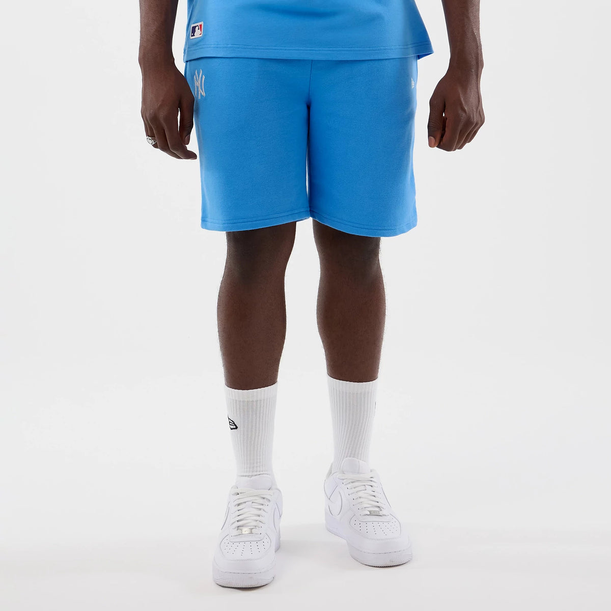 New Era New York Yankees MLB League Essential Pastel Blue Shorts, front view showing the embroidered white New York Yankees logo on the left thigh, styled with a matching blue top, white socks, and white trainers on a model.