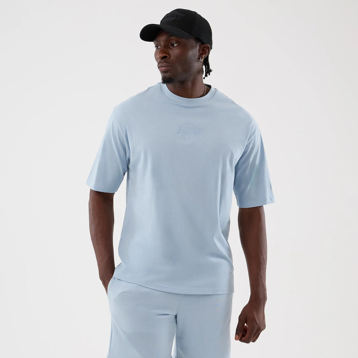 New Era LA Lakers NBA League Essential Pastel Blue Oversized T-Shirt, front view on a model with matching shorts and a black cap, featuring a embossed Lakers logo centred on the chest and short sleeves extending slightly above the elbows.