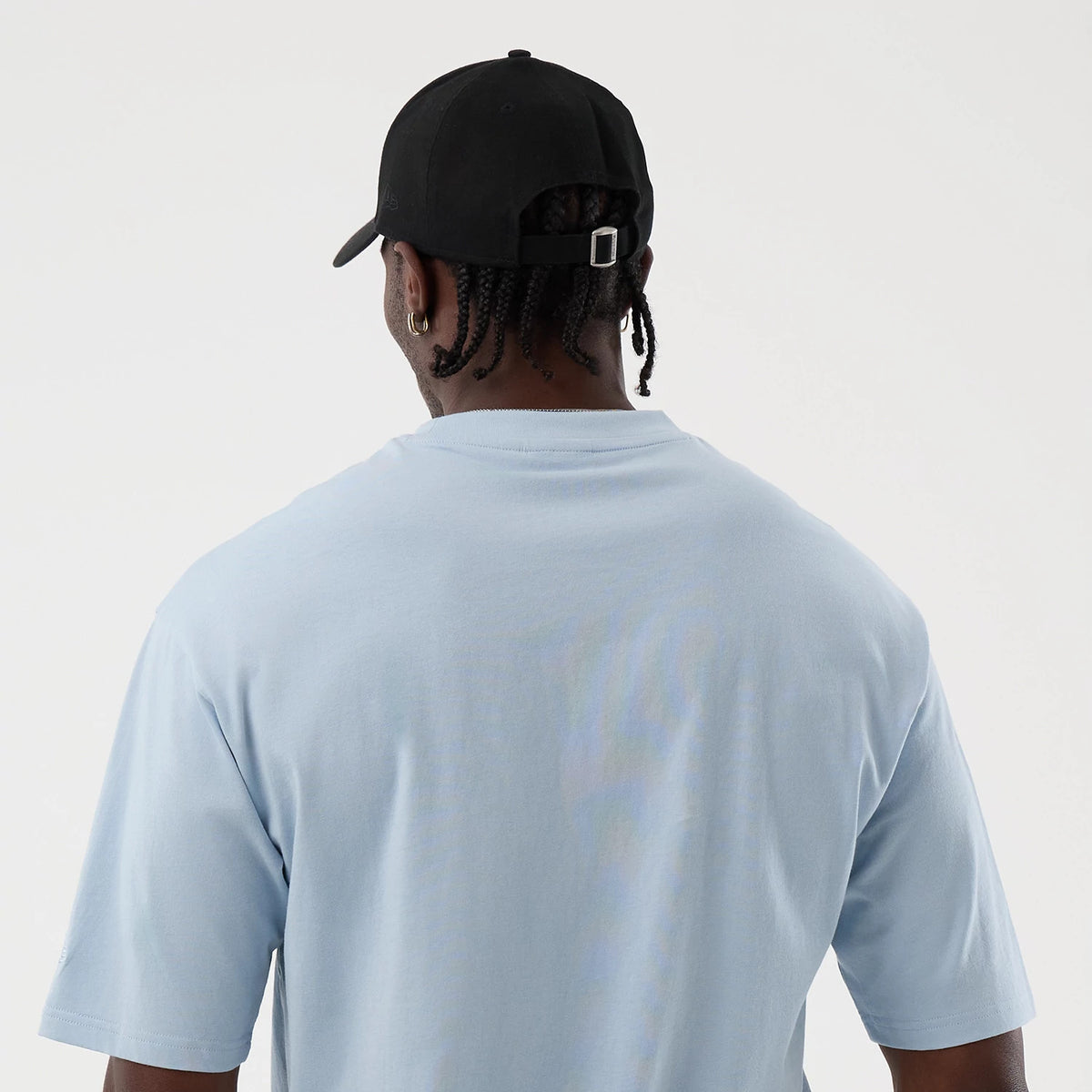 New Era LA Lakers NBA League Essential Pastel Blue Oversized T-Shirt, back view on a model styled with a black cap, showing the shirt's simple design with plain pastel blue fabric and relaxed fit sleeves, highlighting the casual yet sporty look.