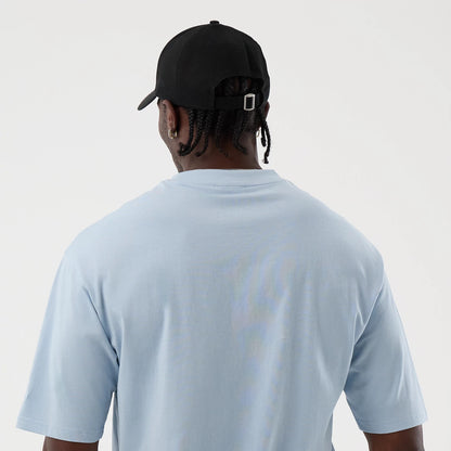 New Era LA Lakers NBA League Essential Pastel Blue Oversized T-Shirt, back view on a model styled with a black cap, showing the shirt's simple design with plain pastel blue fabric and relaxed fit sleeves, highlighting the casual yet sporty look.