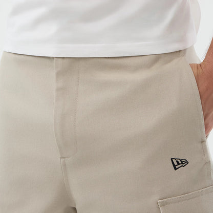 New Era Light Beige Cargo Shorts, front view showing the light beige colour with a simple seam detail, an embroidered New Era logo on the front pocket, and a waistband seen below a white shirt, partially revealing a side pocket.