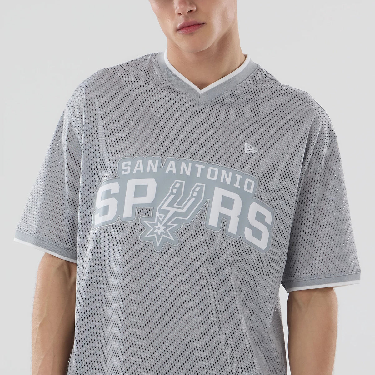 New Era San Antonio Spurs NBA Mesh Grey Oversized T-Shirt, front view on-model displaying a large white printed San Antonio Spurs logo centred across the chest, with a small embroidered New Era logo on the upper right chest and grey V-neck collar.