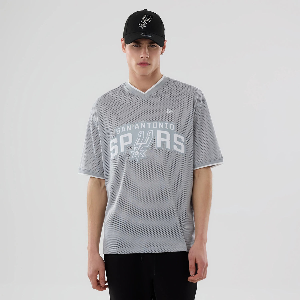 New Era San Antonio Spurs NBA Mesh Grey Oversized T-Shirt, front view on a model wearing a matching black cap, displaying the large white Spurs logo and text across the chest, and a small embroidered New Era logo near the left shoulder.