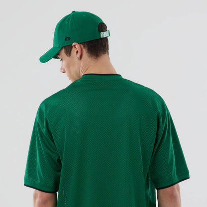 New Era Boston Celtics NBA Mesh Green Oversized T-Shirt, back view on a model with a matching green cap, featuring a plain green mesh design, contrast black ribbed neckline, and black stripe on the sleeves.