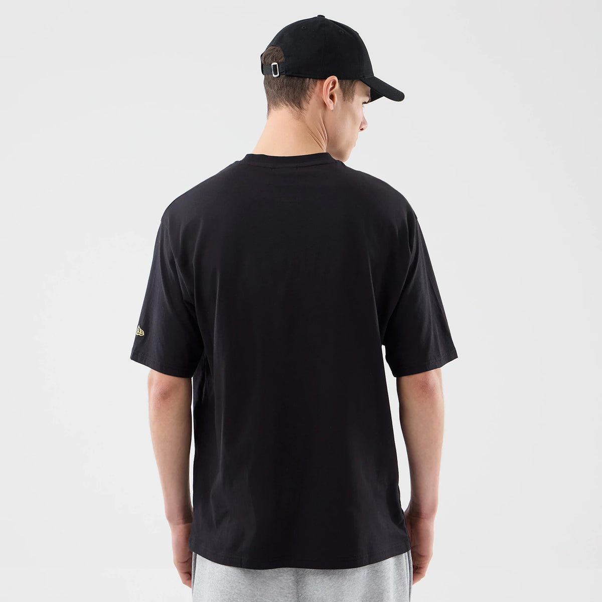 New Era Sticker Porto Black Oversized T-Shirt, back view on a model wearing a black cap, showcasing a plain black back with a embroidered New Era logo in gold on the left sleeve and a relaxed fit over grey trousers.