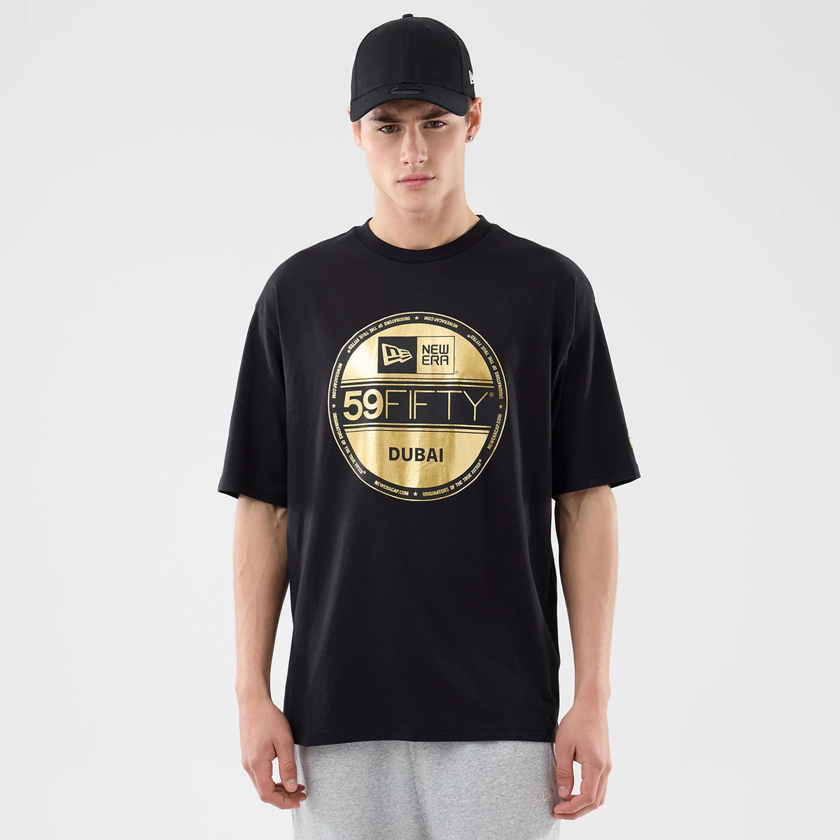 New Era Sticker Dubai Black Oversized T-Shirt, front view on-model showing a gold circular graphic with the New Era logo, 59FIFTY and Dubai text centred on the black oversized tee, paired with a plain black cap.