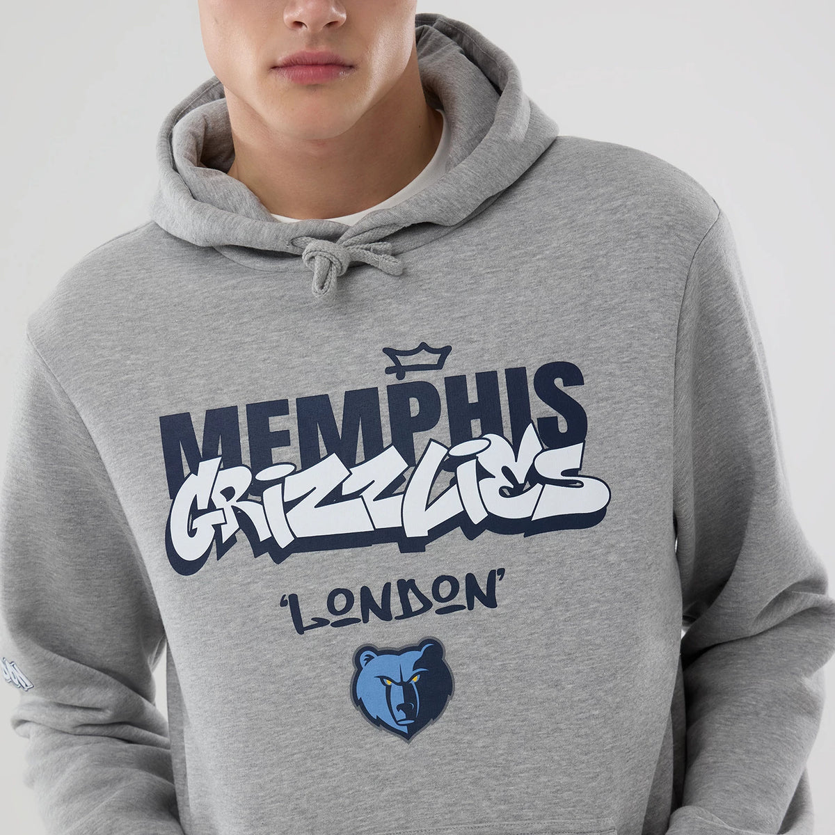 New Era Memphis Grizzlies NBA London Game 2026 Grey Pullover Hoodie, front view on a model, showcasing navy and white printed Memphis Grizzlies lettering across the chest, an additional crown graphic above.