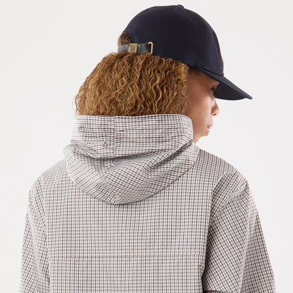 Womens New Era Check Beige Oversized Windbreaker Jacket, back view on a model showing the beige and grey check pattern, the hood hanging behind the neck, and a glimpse of a navy cap with an embroidered New Era logo on the strap.