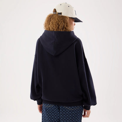 New Era New York Yankees Womens MLB Navy Oversized Pullover Hoodie, back view on a model, showcasing the loose-fitting navy hooded sweatshirt with relaxed sleeves and hem, styled with patterned trousers and a light-coloured cap with eyelets.