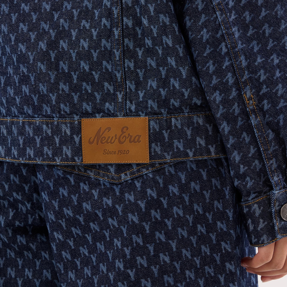 New Era New York Yankees Womens MLB Denim Navy Jacket, close-up showing an all-over N pattern on the navy denim fabric, a brown patch with a debossed New Era logo above the hem, and button detail on the cuff.