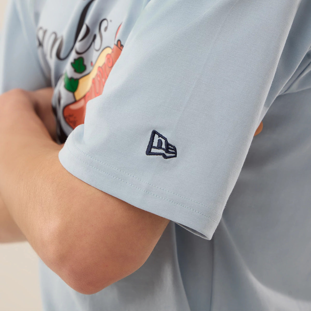 New Era New York Yankees MLB Food Graphic Pastel Blue Oversized T-Shirt, side view featuring an embroidered New Era logo on the left sleeve, an arm crossed in front and a hot dog and Yankees script graphic partially on the chest.