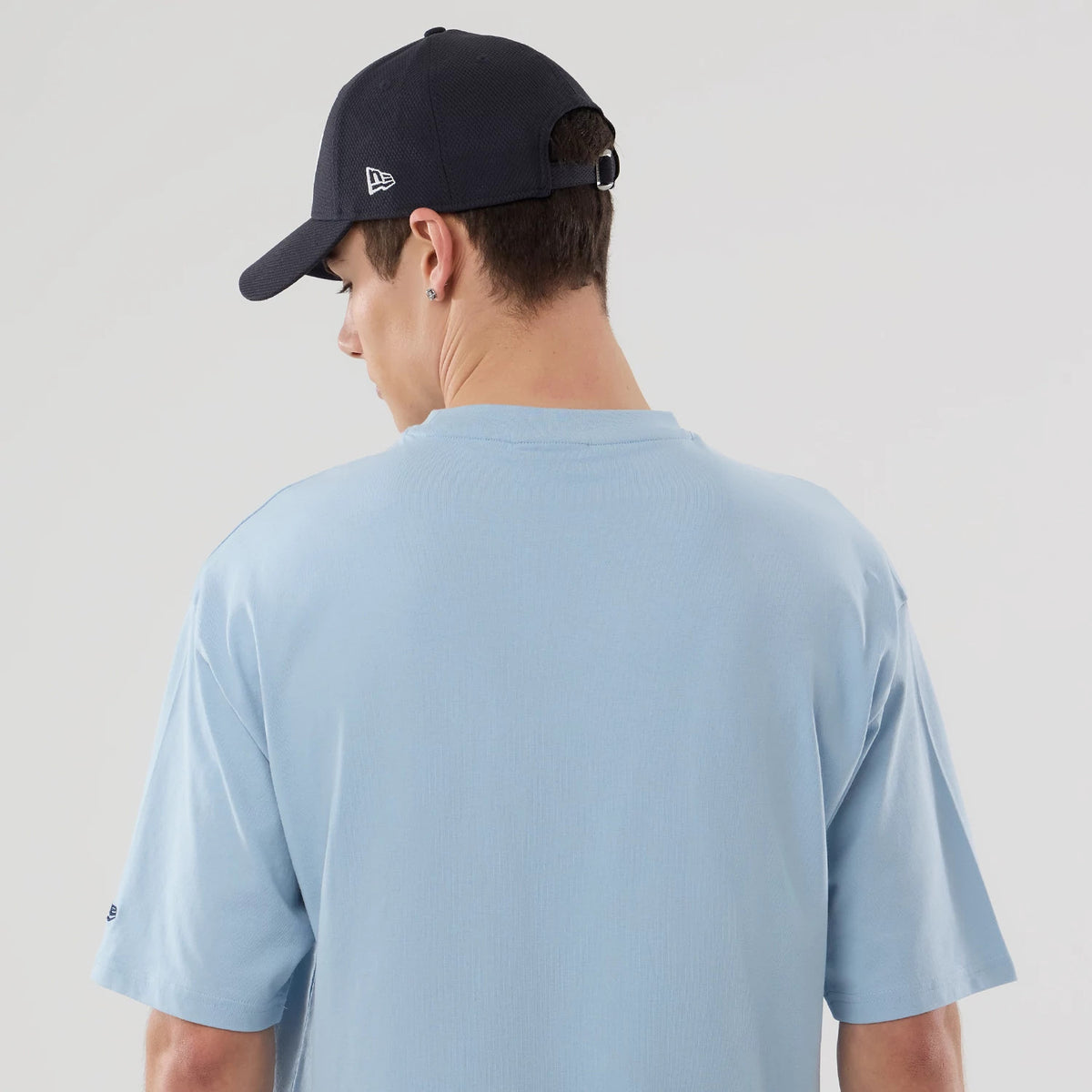 New Era New York Yankees MLB Food Graphic Pastel Blue Oversized T-Shirt, back view on a model wearing a dark cap, showing the plain pastel blue fabric with relaxed short sleeves and an embroidered New Era logo on the left sleeve near the cuff.