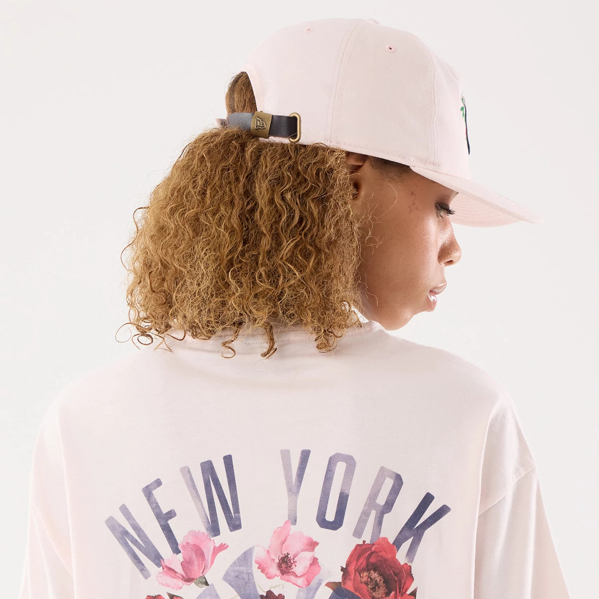 New Era New York Yankees Womens MLB Graphic Pastel Pink T-Shirt, back view on a model featuring large New York lettering above pink floral graphics, paired with a pastel pink cap showing an embroidered logo and adjustable strap at the rear.