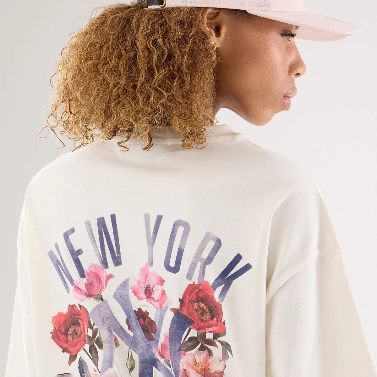 New Era New York Yankees Womens MLB Graphic Open Cream T-Shirt, back view on a model with a light pink cap, showing New York lettering above the Yankees logo adorned with red and pink flowers, giving a and feminine touch.