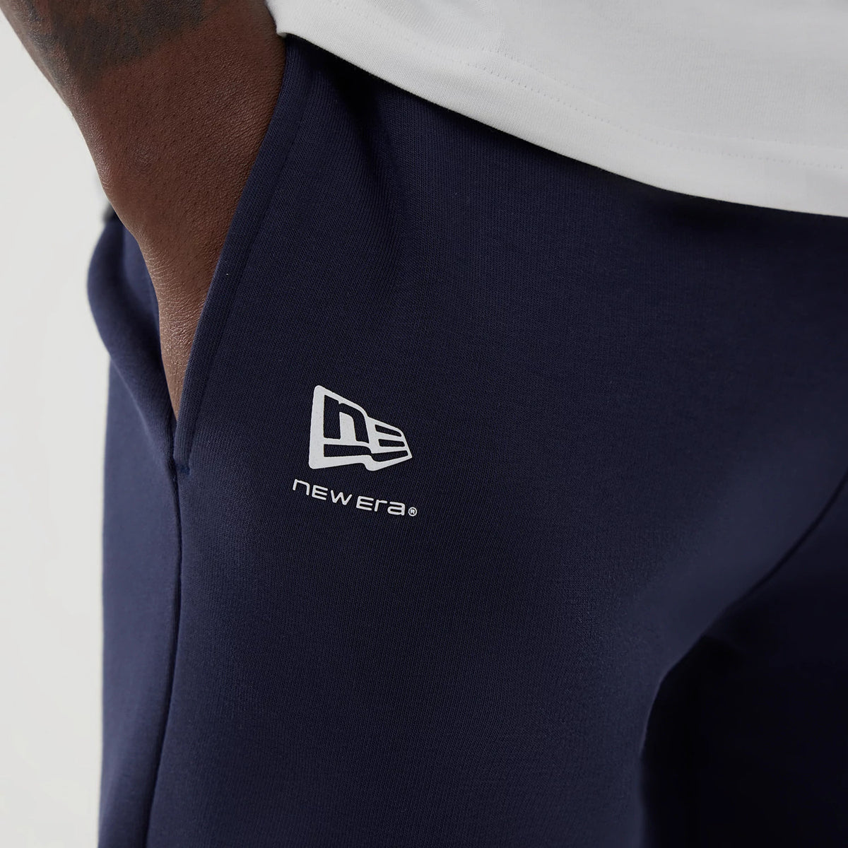 New Era Williams Navy Shorts, on-model close-up showing dark blue shorts with a contrasting white printed New Era logo on the left thigh, styled with a white top, side pocket and casual relaxed fit.