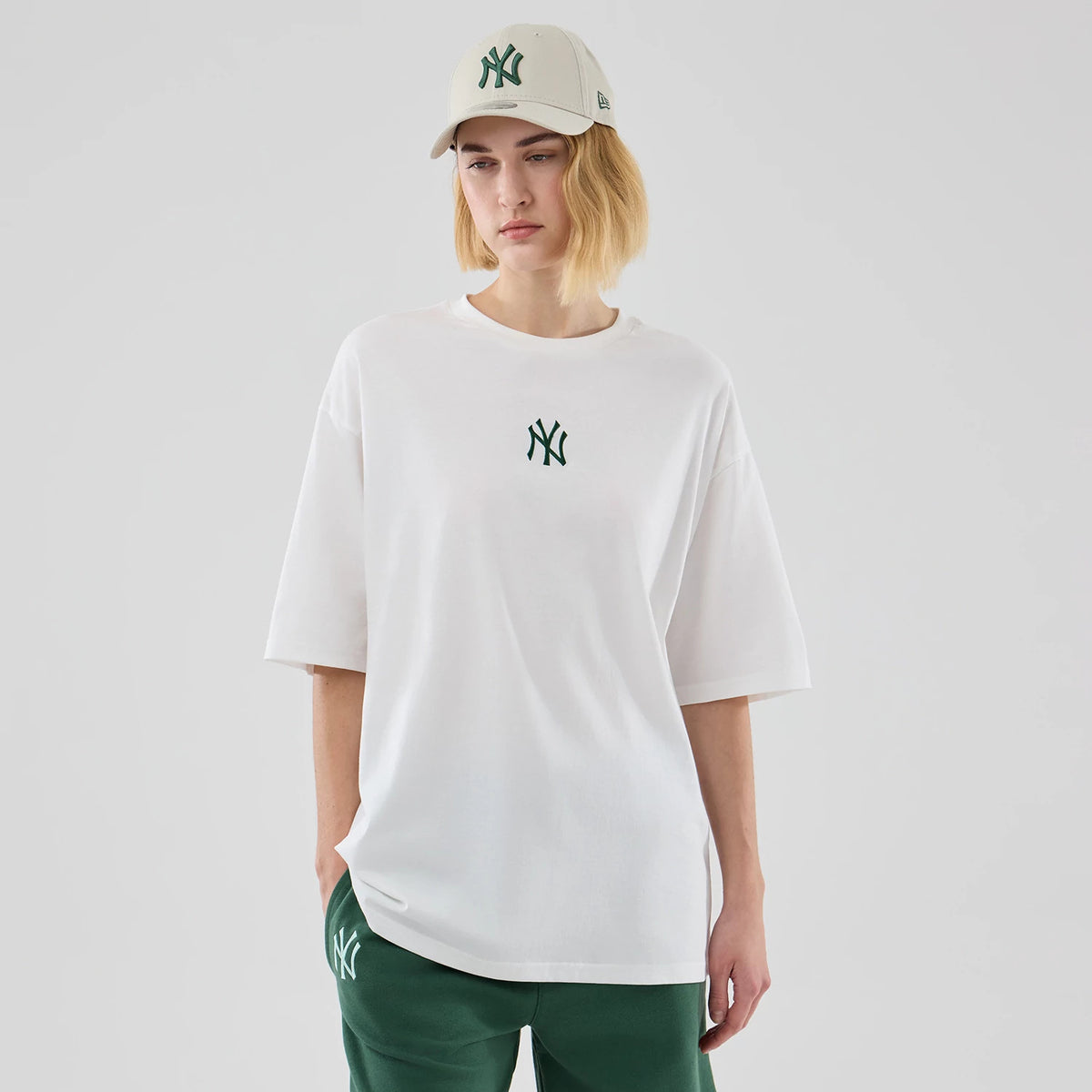 New Era New York Yankees MLB Midi League Essential White Oversized T-Shirt, front view on a model wearing a cap, showing a green embroidered Yankees logo centred on the chest, short sleeves with a wide fit, and styled with matching green trousers.