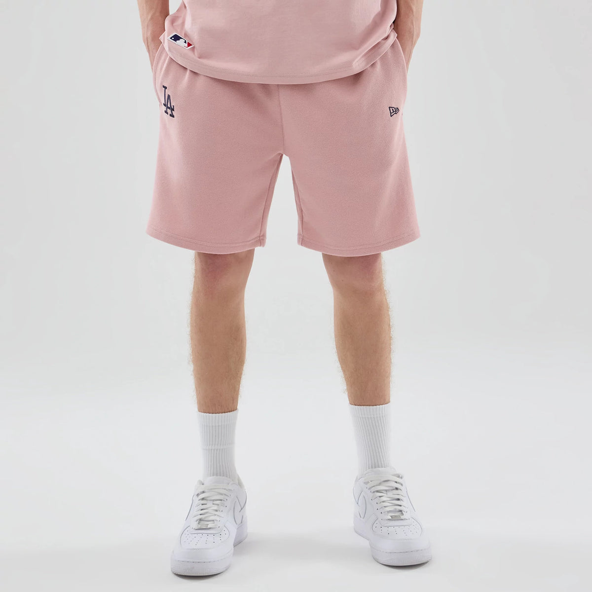 New Era LA Dodgers MLB Midi League Essential Pastel Pink Oversized Shorts, front view on-model showing the pink shorts reaching just above the knee, with a black embroidered LA logo near the left hem and a black embroidered New Era logo on the right.