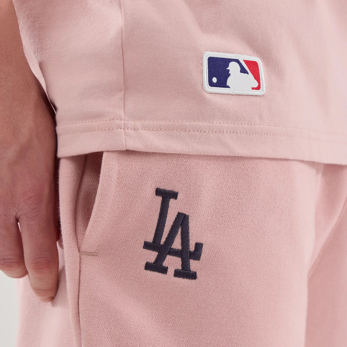 New Era LA Dodgers MLB Midi League Essential Pastel Pink Oversized Shorts, side view showing a black embroidered LA logo on the left thigh, a white embroidered MLB logo above the hem, and a close view of the pocket detail.