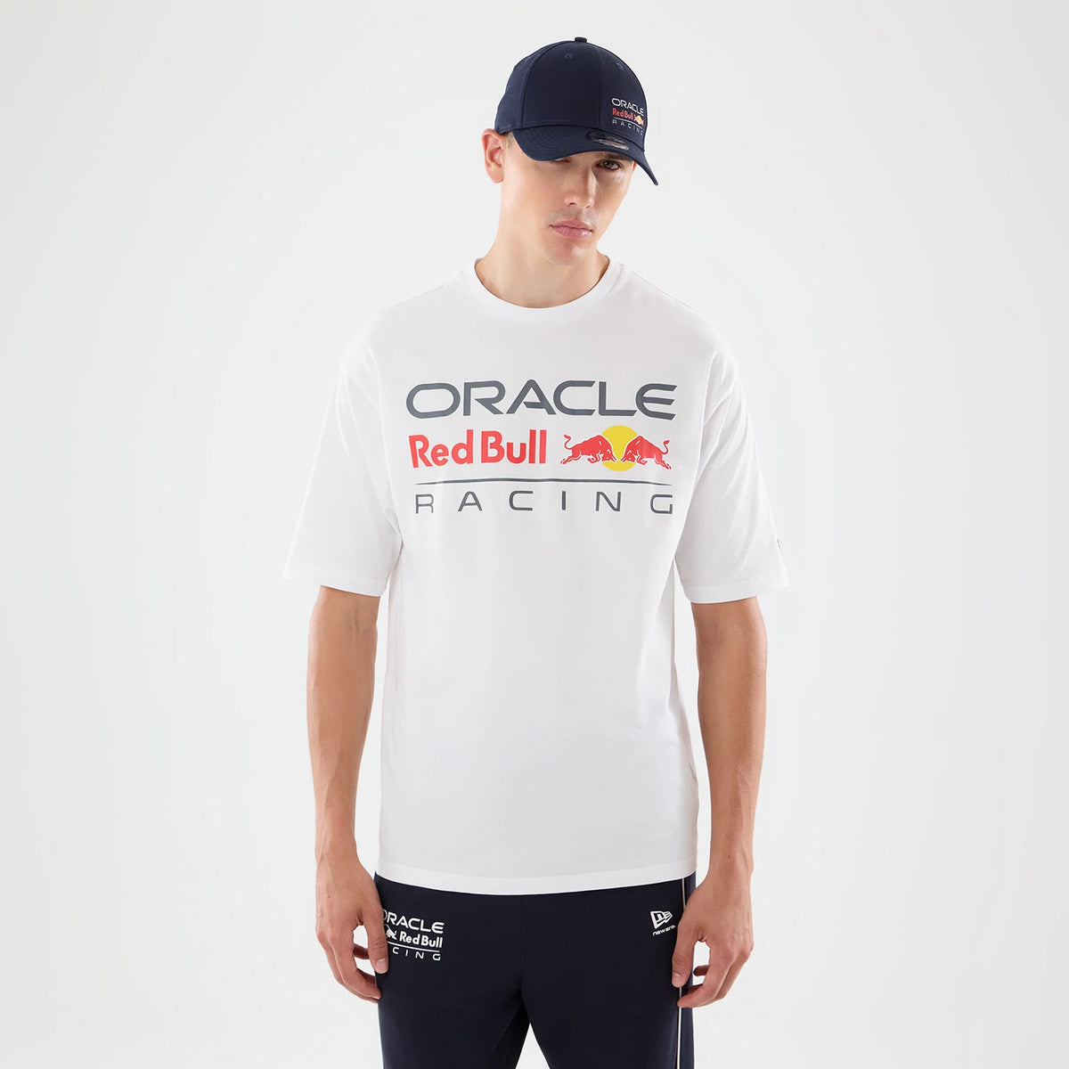 New Era Red Bull Racing Graphic White Oversized T-Shirt, front view on-model showing Oracle and Red Bull Racing lettering with the red bull graphics across the chest, paired with navy trousers featuring embroidered team branding on the leg.
