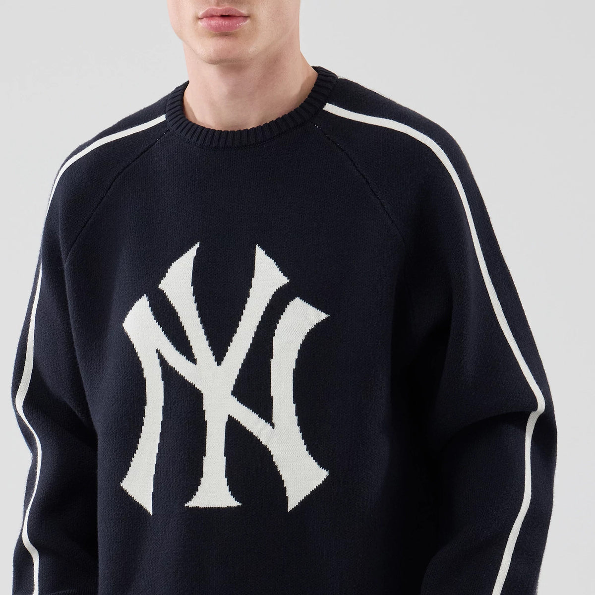 New Era New York Yankees MLB Knit Navy Knitted Sweater, front view on a model featuring a large white NY logo knitted onto the chest, ribbed crew neck collar, and white stripe running down the sleeves.