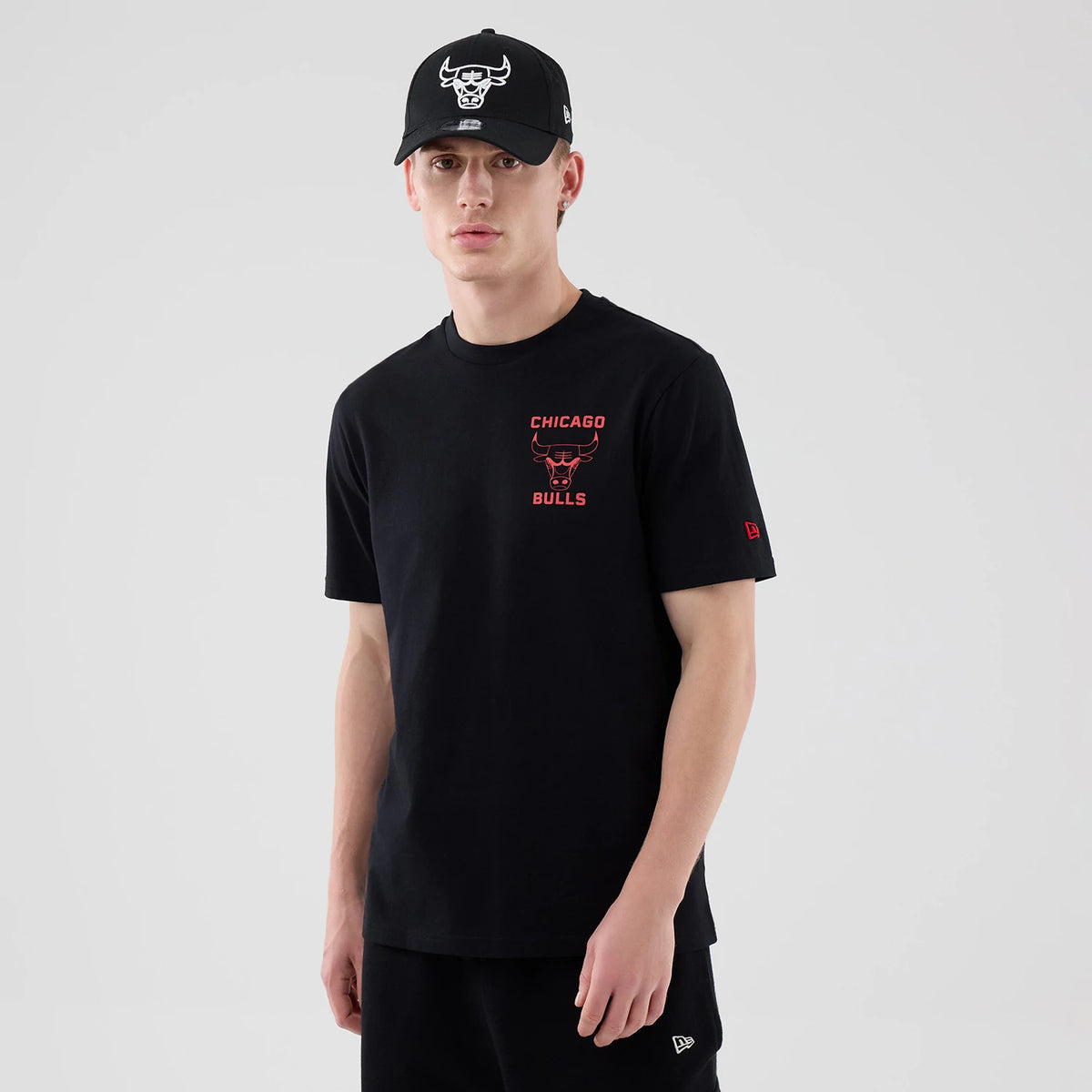 New Era Chicago Bulls NBA Court Graphic Black T-Shirt, front view on-model showing the red printed team logo and lettering on the left chest, paired with a black cap featuring an embroidered white bull logo and New Era flag on the side.