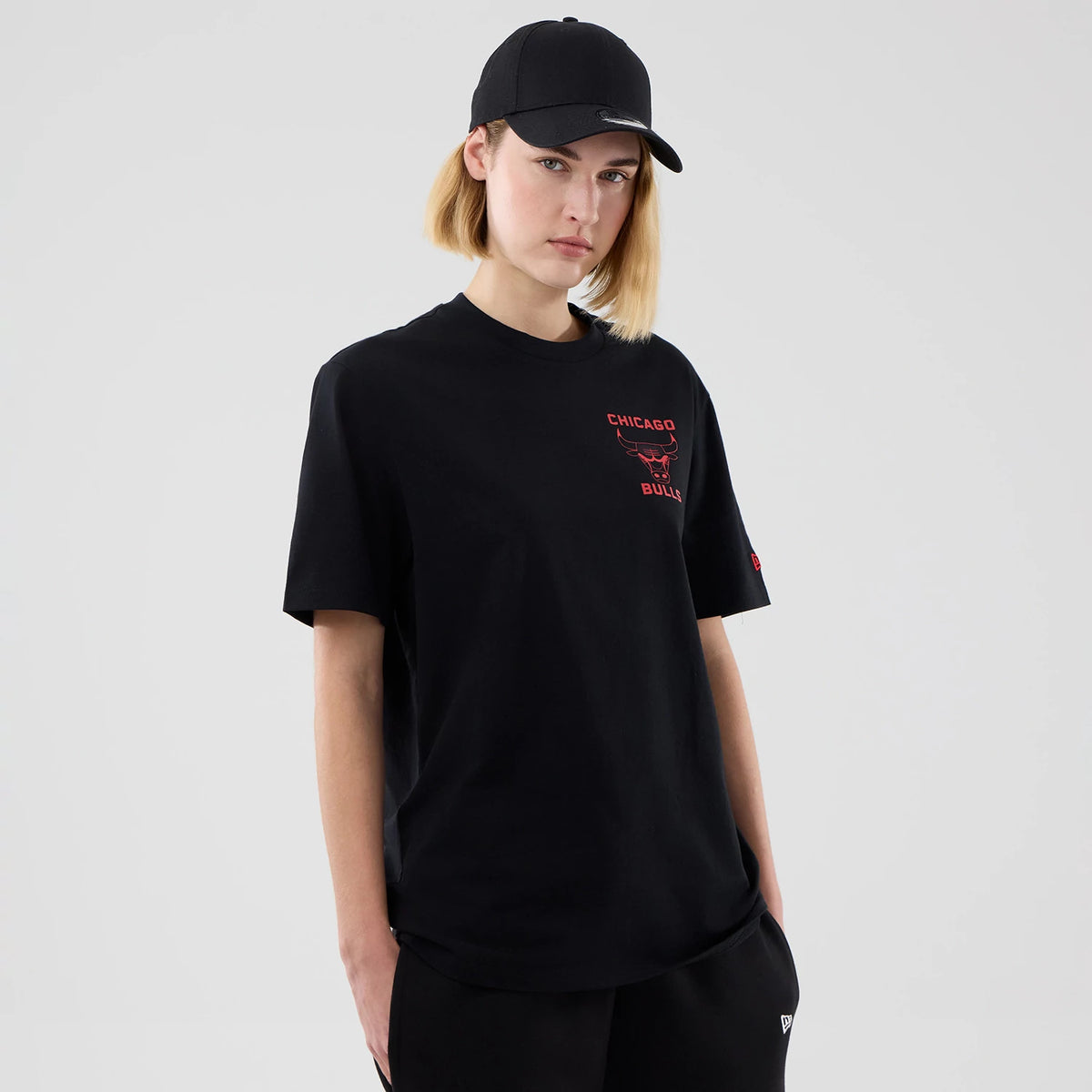 New Era Chicago Bulls NBA Court Graphic Black T-Shirt, front view on-model showing the black short-sleeved tee with a red printed Chicago Bulls logo and text on the left chest, paired with black trousers and a black cap.