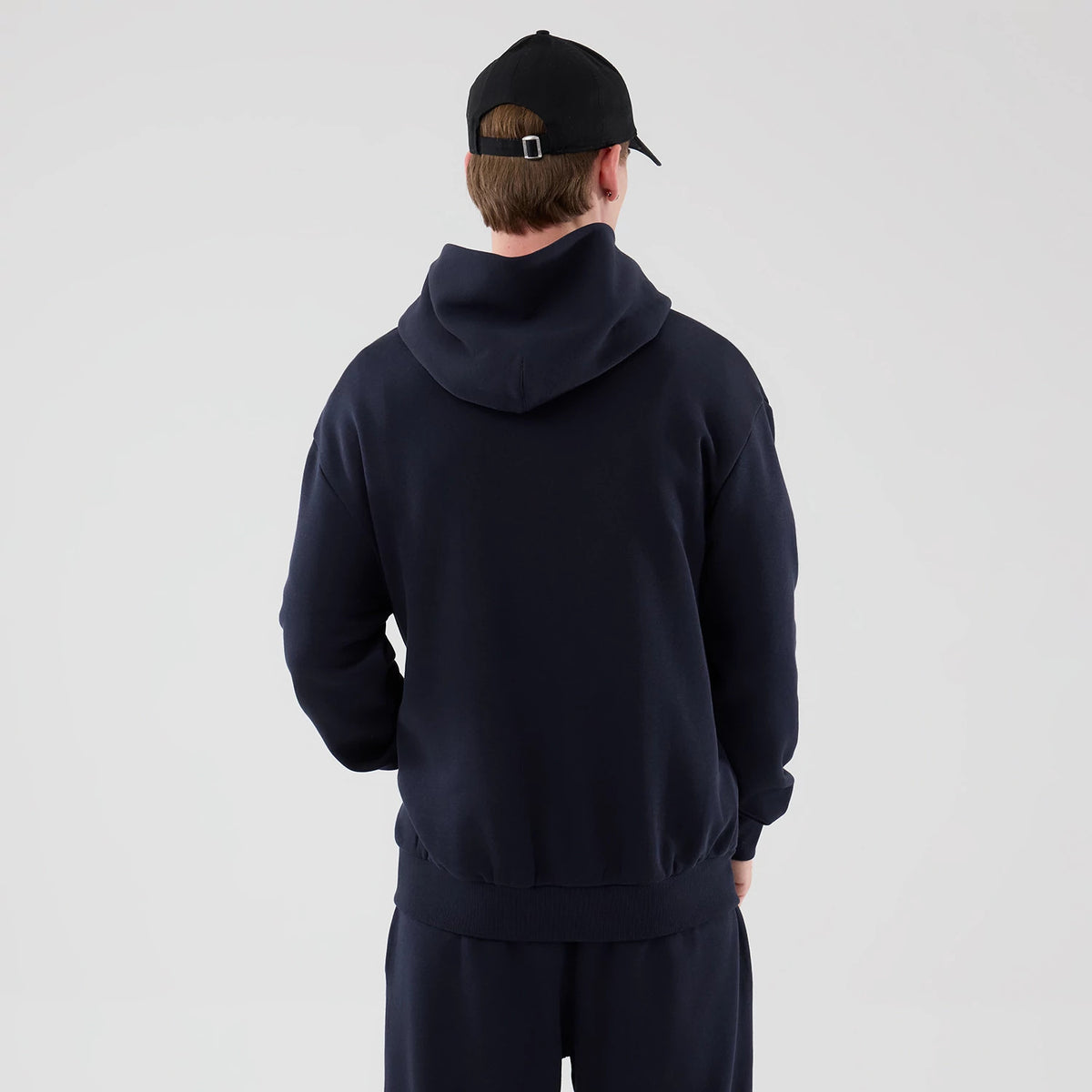 New Era LA Lakers NBA Core Plus Navy Pullover Hoodie, back view on a model paired with matching navy trousers and a black cap, highlighting the oversized hood and relaxed fit with ribbed cuffs and hem for casual styling.