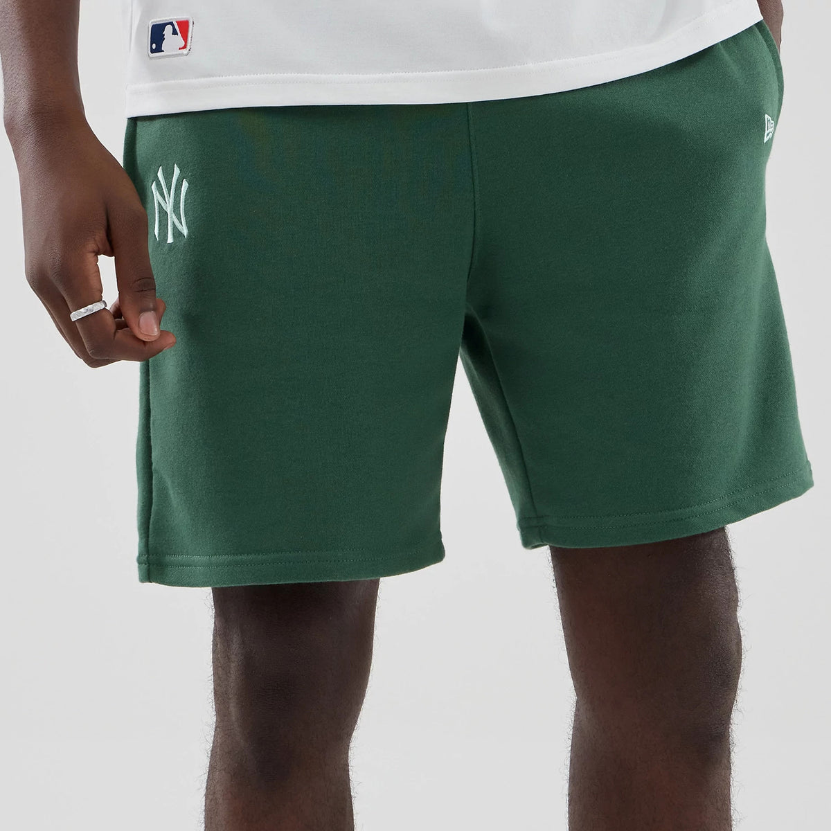New Era New York Yankees MLB Midi League Essential Dark Green Oversized Shorts, front view showing the white embroidered NY logo on the left side, the New Era flag above the hem on the right, and the shorts styled with a white top.