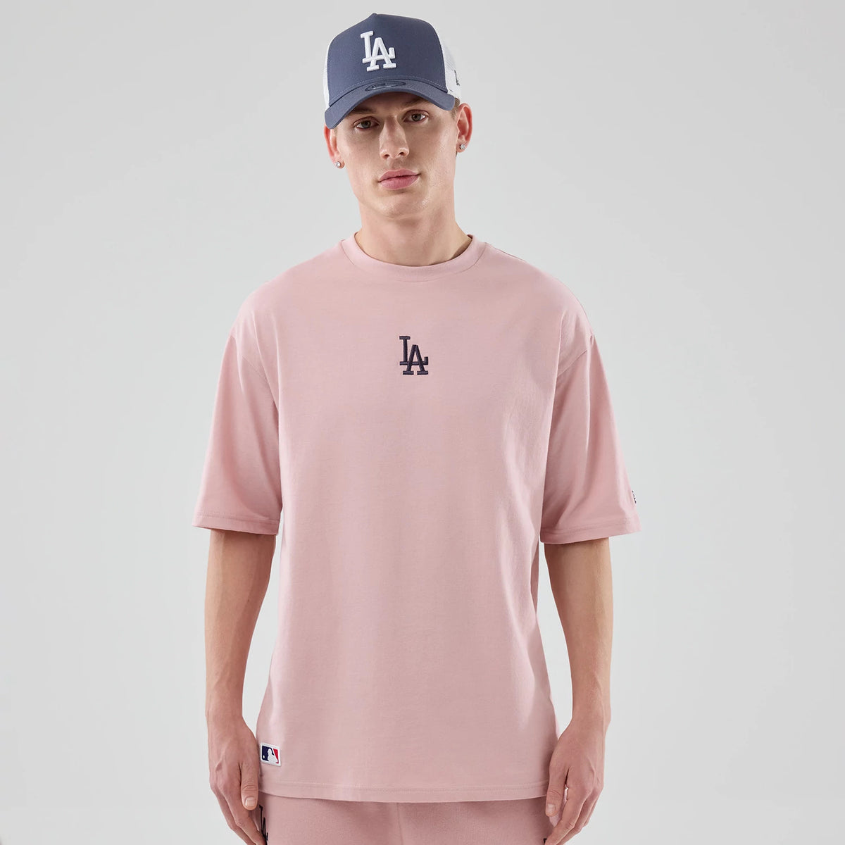New Era LA Dodgers MLB Midi League Essential Pastel Pink Oversized T-Shirt, front view on a model showing the black embroidered LA logo centred on the chest, with loose short sleeves and a small MLB logo patch near the lower hem.