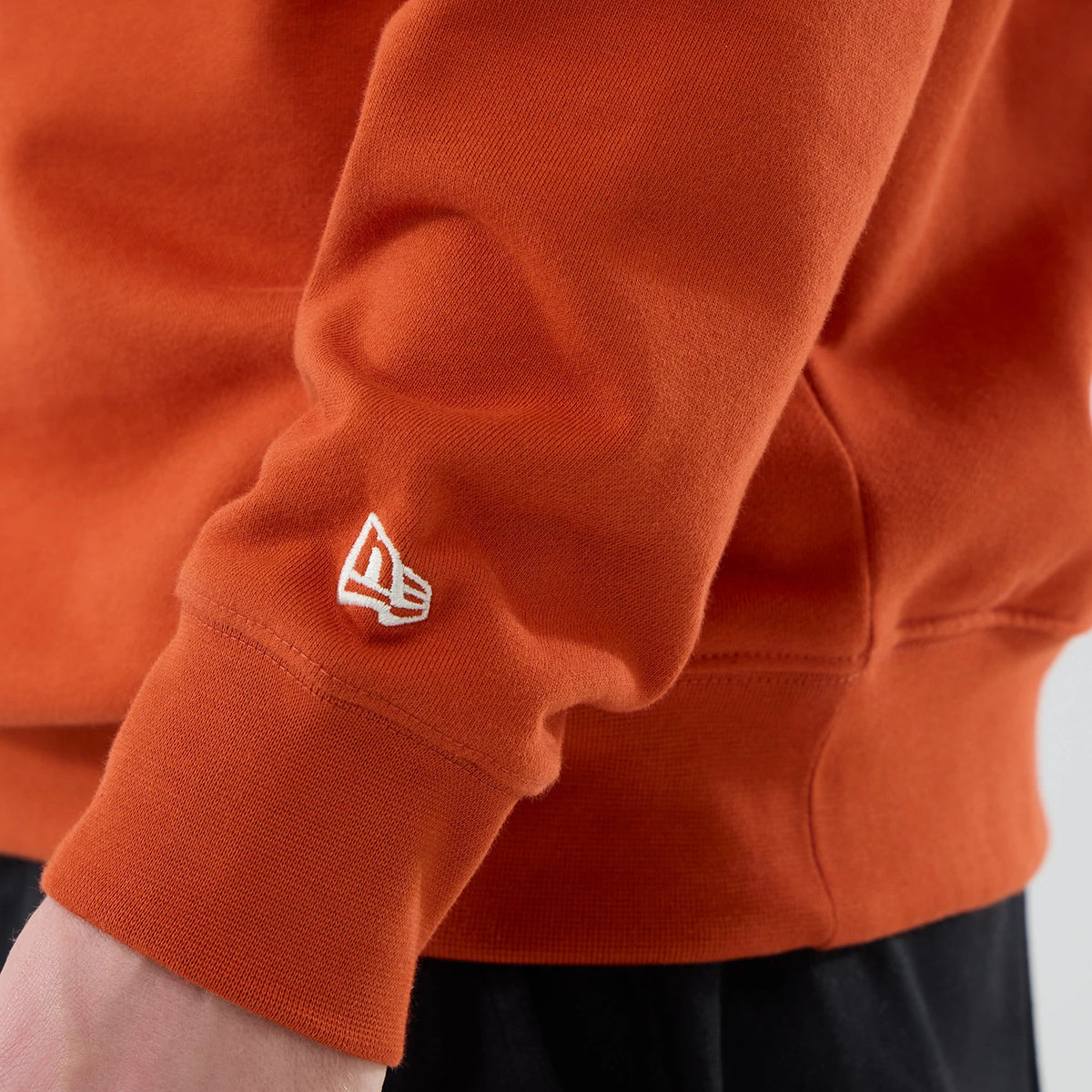 New Era New York Yankees MLB Arch Graphic Orange Oversized Crew Neck Sweater, close-up view showing the white embroidered New Era flag logo on the left sleeve cuff, with ribbed and a part of the hem above black trousers.