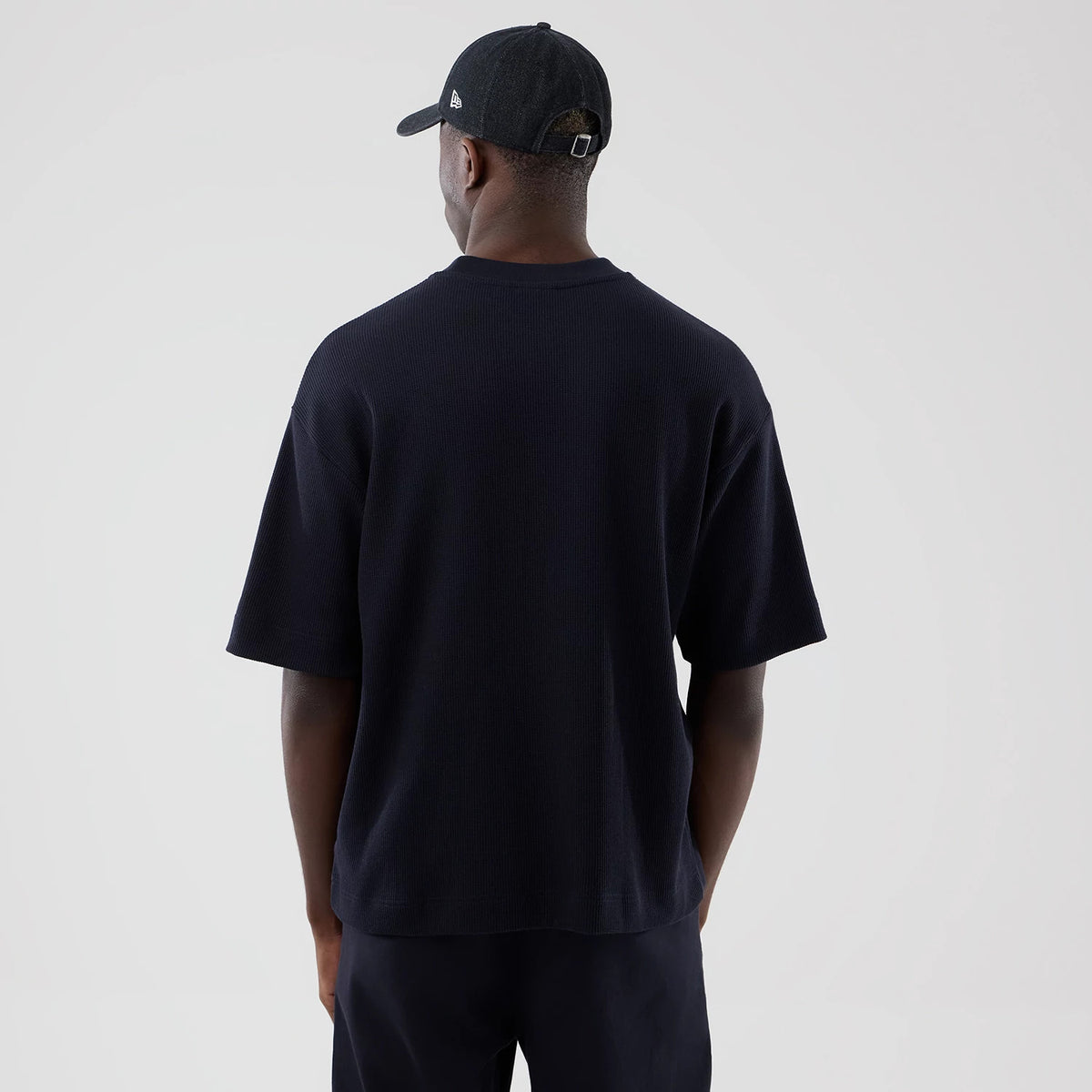 New Era Waffle Navy Boxy T-Shirt, back view on a model wearing matching navy trousers and a black cap, highlighting the loose fit and wide sleeves, with a plain navy texture and a drop hem for a casual look.