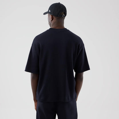 New Era Waffle Navy Boxy T-Shirt, back view on a model wearing matching navy trousers and a black cap, highlighting the loose fit and wide sleeves, with a plain navy texture and a drop hem for a casual look.