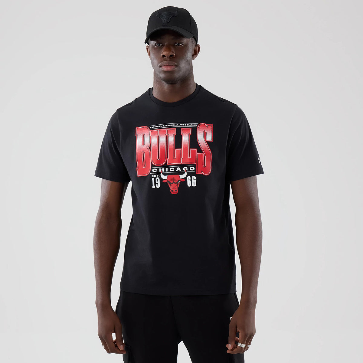 New Era Chicago Bulls NBA Arch Graphic Black T-Shirt, front view on a model with a black cap, showing a large red and white 'BULLS' print across the chest, a small Bulls logo beneath, and the number '1966' on either side of the logo.