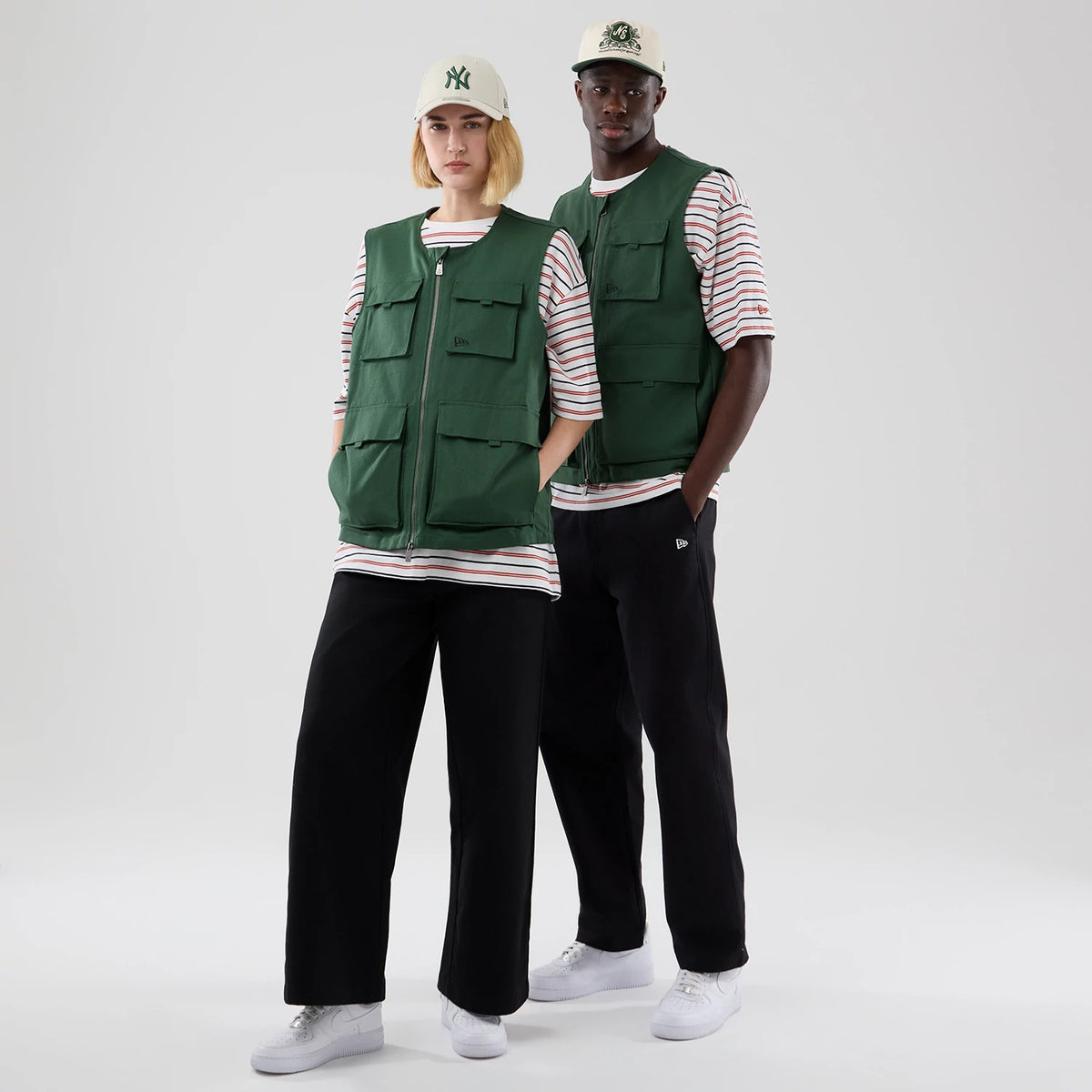 New Era Dark Green Utility Vest, on-model view with two people wearing white caps, showcasing front zip closure, large front pockets, and a dark green colour, layered over striped shirts and paired with black trousers and white trainers.