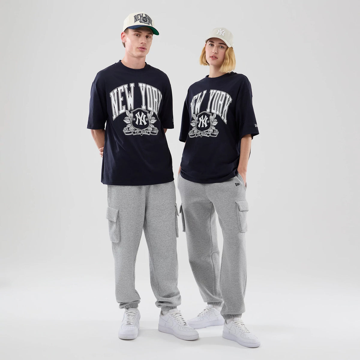 New Era New York Yankees MLB Arch Graphic Navy Oversized T-Shirt, front view on-model showing large white New York lettering with the Yankees logo in the centre, complemented by grey cargo trousers and white trainers, with caps adding a casual style.