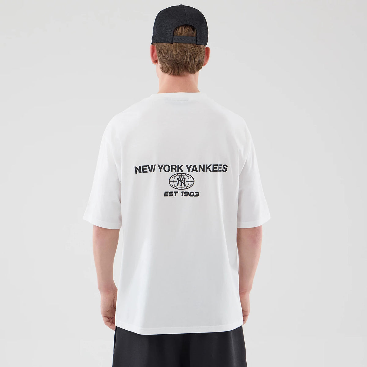 New Era New York Yankees MLB Logo Graphic White Oversized T-Shirt, back view on a model wearing a dark cap, featuring black printed lettering NEW YORK YANKEES above a circular Yankees logo and EST 1903 centred across the upper back.