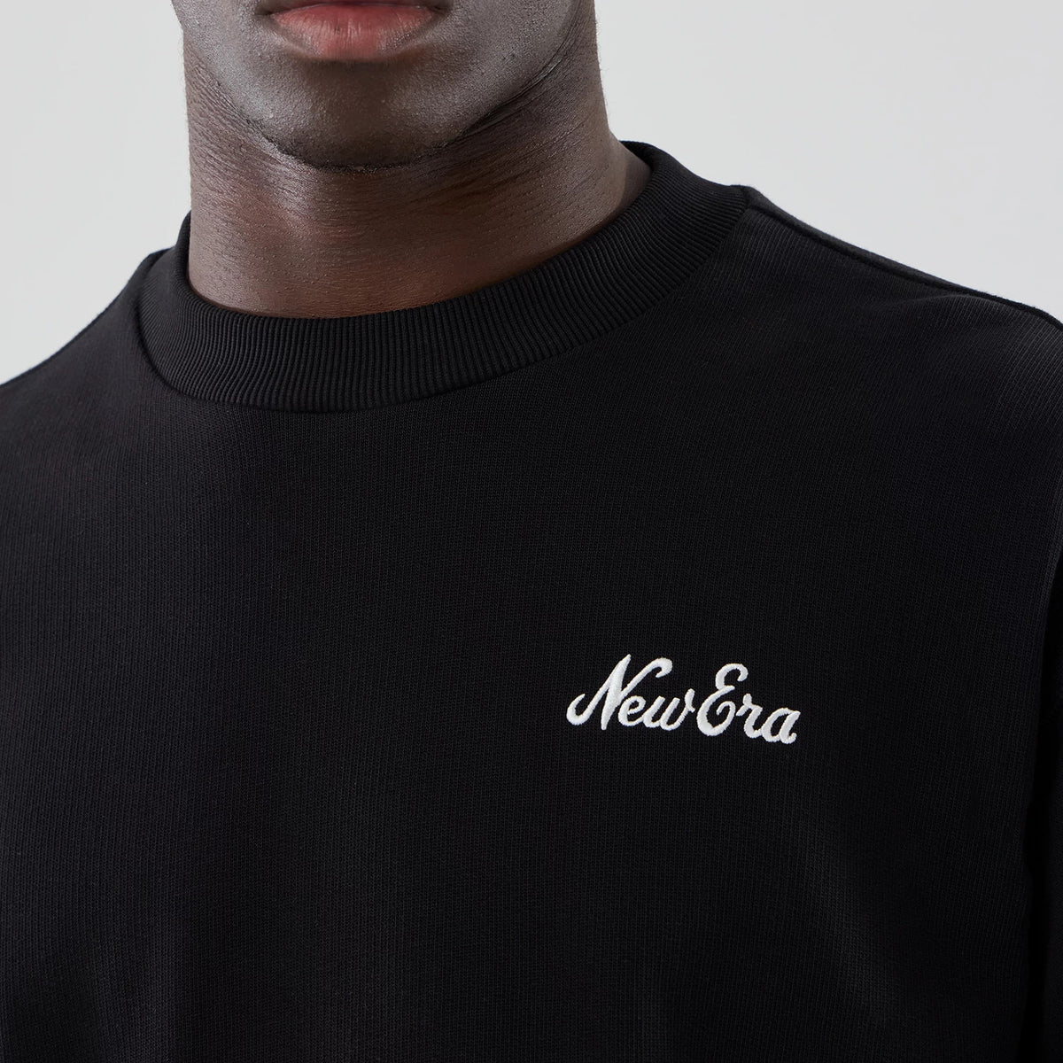 New Era Black Oversized Crew Neck Sweater, close-up on a model showing the ribbed crew neck and the white embroidered New Era script logo on the upper left chest of the black fabric.