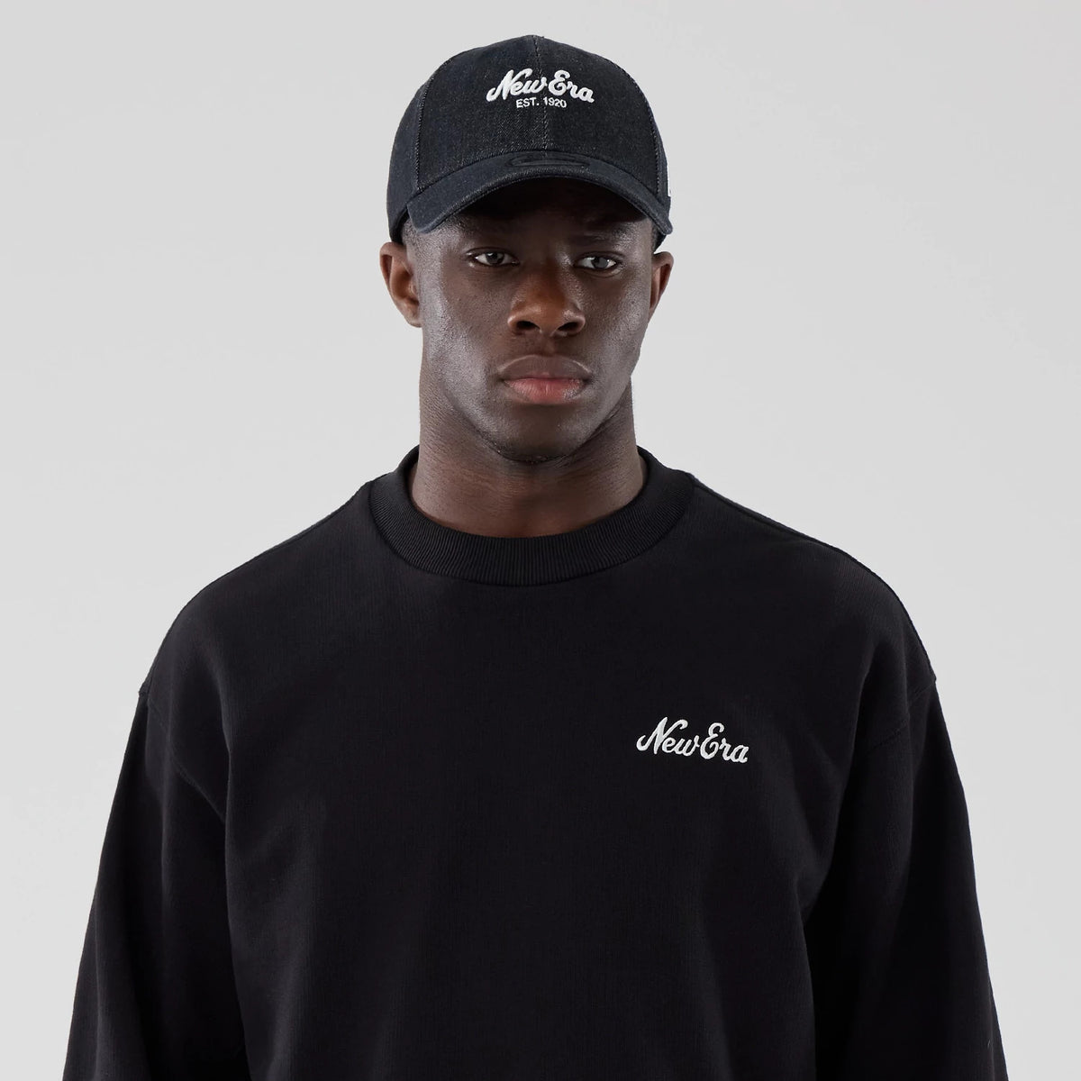 New Era Black Oversized Crew Neck Sweater, front view on a model wearing a matching black cap, featuring the New Era logo embroidered in white on the left chest and showcasing the relaxed fit and ribbed neckline.