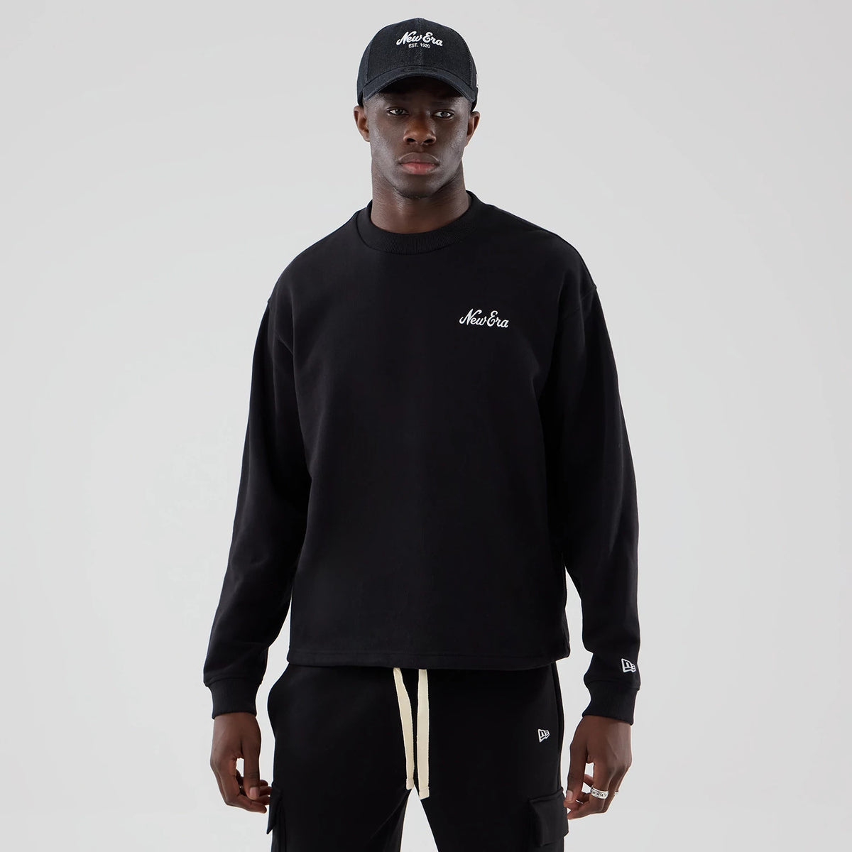 New Era Black Oversized Crew Neck Sweater, front view on a model wearing a matching black cap, featuring a white embroidered New Era logo on the left chest and another embroidered logo on the left sleeve, with ribbed cuffs.