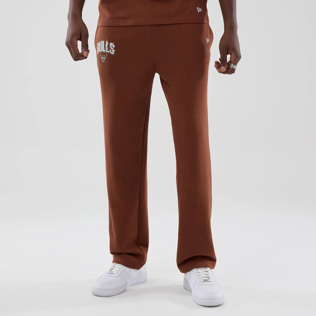 New Era Chicago Bulls NBA Core Plus Brown Straight Leg Joggers, front view on a model wearing white trainers, showing the Bulls emblem in white on the right thigh and the New Era flag logo on the left side by the pocket.