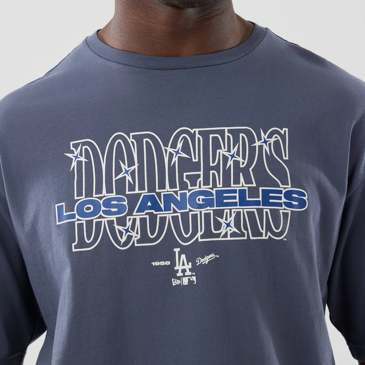 New Era LA Dodgers MLB Graphic Grey Oversized T-Shirt, front view showing Dodgers lettering with star details across the chest, Los Angeles text beneath in bright blue, and embroidered LA team logos at the bottom centre, styled casually on a person.