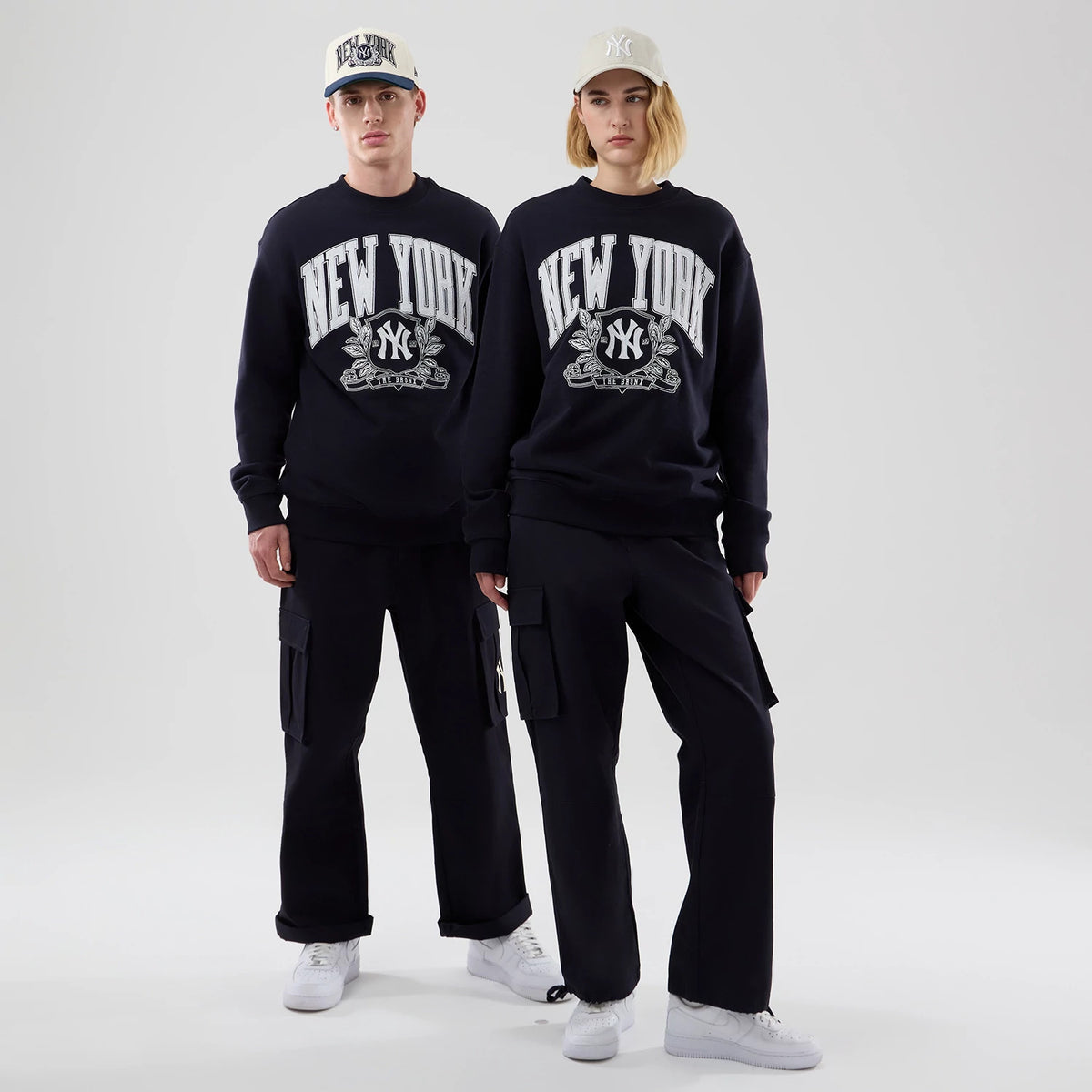 New Era New York Yankees MLB Arch Graphic Navy Oversized Crew Neck Sweater, front view on models with matching caps, displaying a large white New York text above the Yankees logo on the navy fabric, paired with black cargo trousers.
