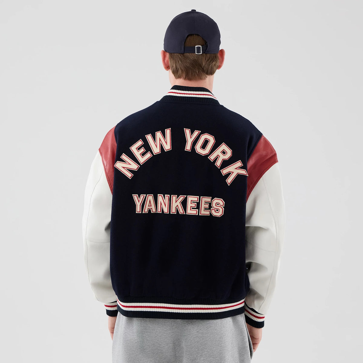 New Era New York Yankees MLB Varsity Navy Jacket, back view on a model featuring large raised New York Yankees lettering in white and red across the shoulders and back, with red sleeve accents and white sleeves, paired with a dark baseball cap.