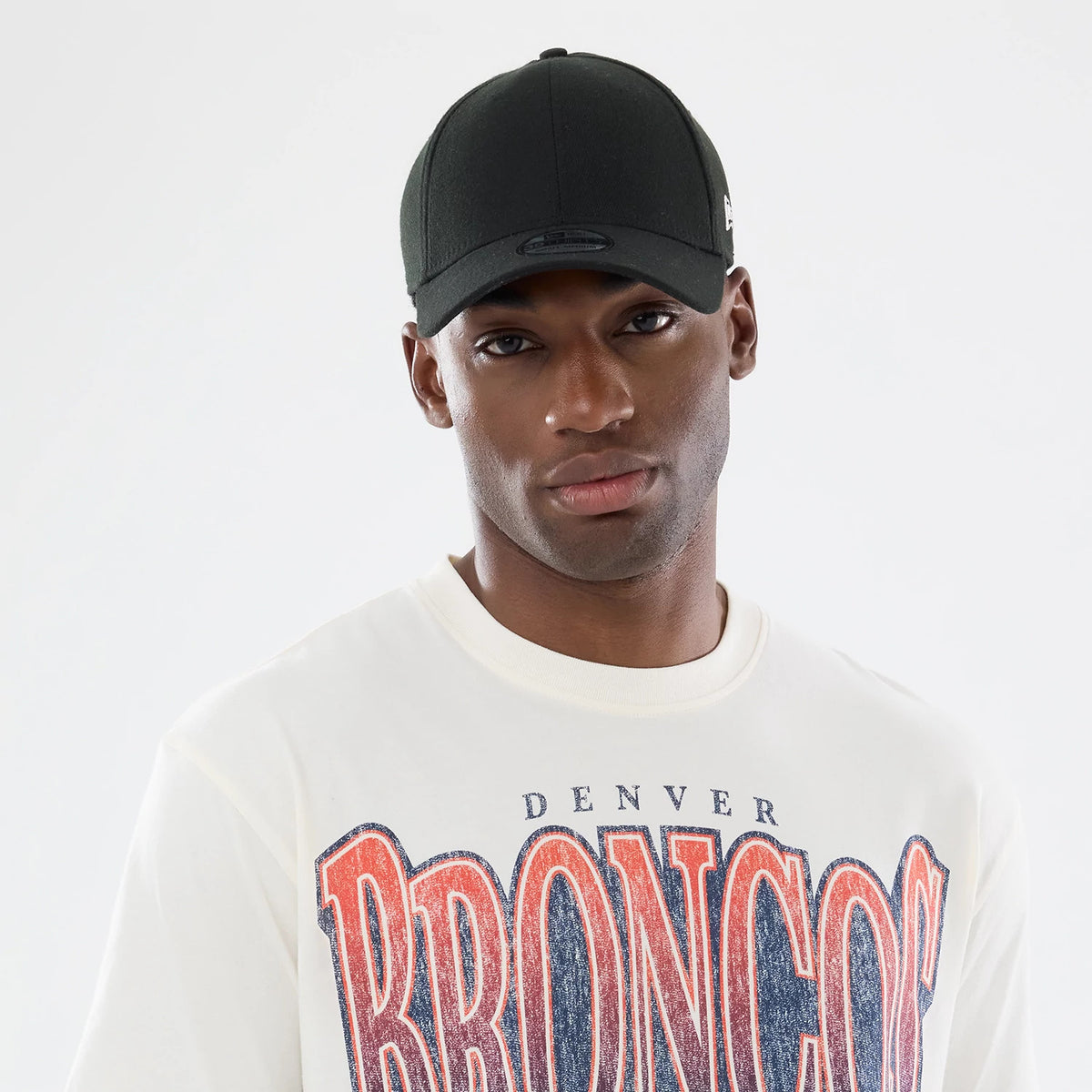 New Era Denver Broncos NFL Sport Classic Cream T-Shirt, front view on a model paired with a black cap, featuring large distressed orange lettering spelling BRONCOS on the front center of the cream shirt with smaller DENVER text above.