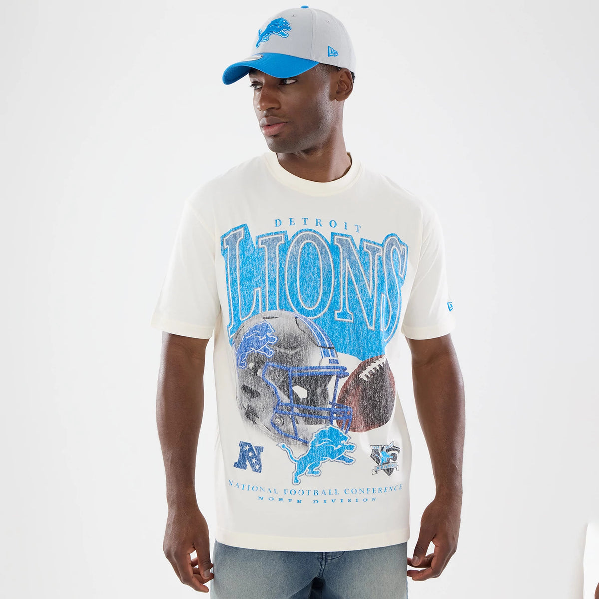 New Era Detroit Lions NFL Sport Classic Cream T-Shirt, front view on a model with blue jeans and a two-tone cap, featuring large LIONS lettering in blue, a detailed graphic of a helmet and football, and a small embroidered New Era logo on the sleeve.