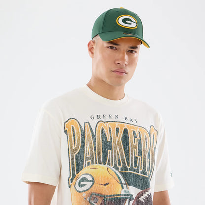 New Era Green Bay Packers NFL Sport Classic Cream T-Shirt, front view on a model wearing a matching green cap, featuring Packers lettering across the chest and a graphic of a football helmet, with green Bay text at the top.