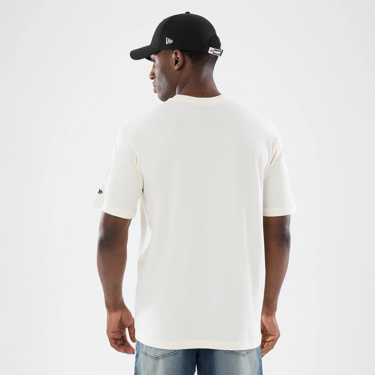 New Era Las Vegas Raiders NFL Sport Classic Cream T-Shirt, back view on a model wearing jeans and a black cap, showing no graphics on the cream back, an embroidered New Era logo on the left sleeve, and a relaxed fit.
