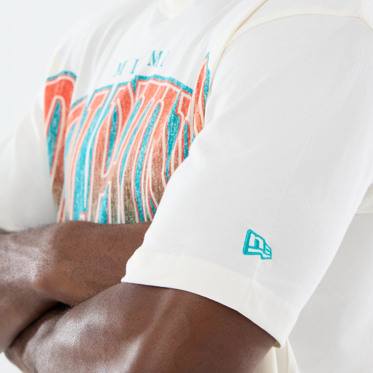 New Era Miami Dolphins NFL Sport Classic Cream T-Shirt, front view on a model with arms crossed, highlighting the Miami Dolphins lettering in vivid teal and orange across the chest and an embroidered New Era logo on the left sleeve.