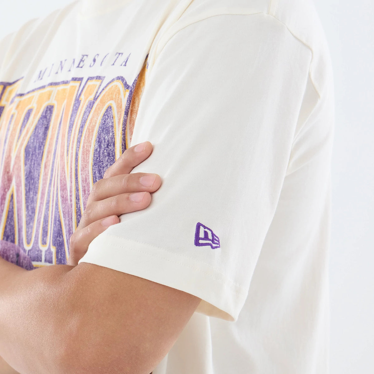 New Era Minnesota Vikings NFL Sport Classic Cream T-Shirt, three-quarter view showing the large VIKINGS lettering across the chest in purple and gold, with a purple embroidered New Era logo on the sleeve, styled casually on a person.